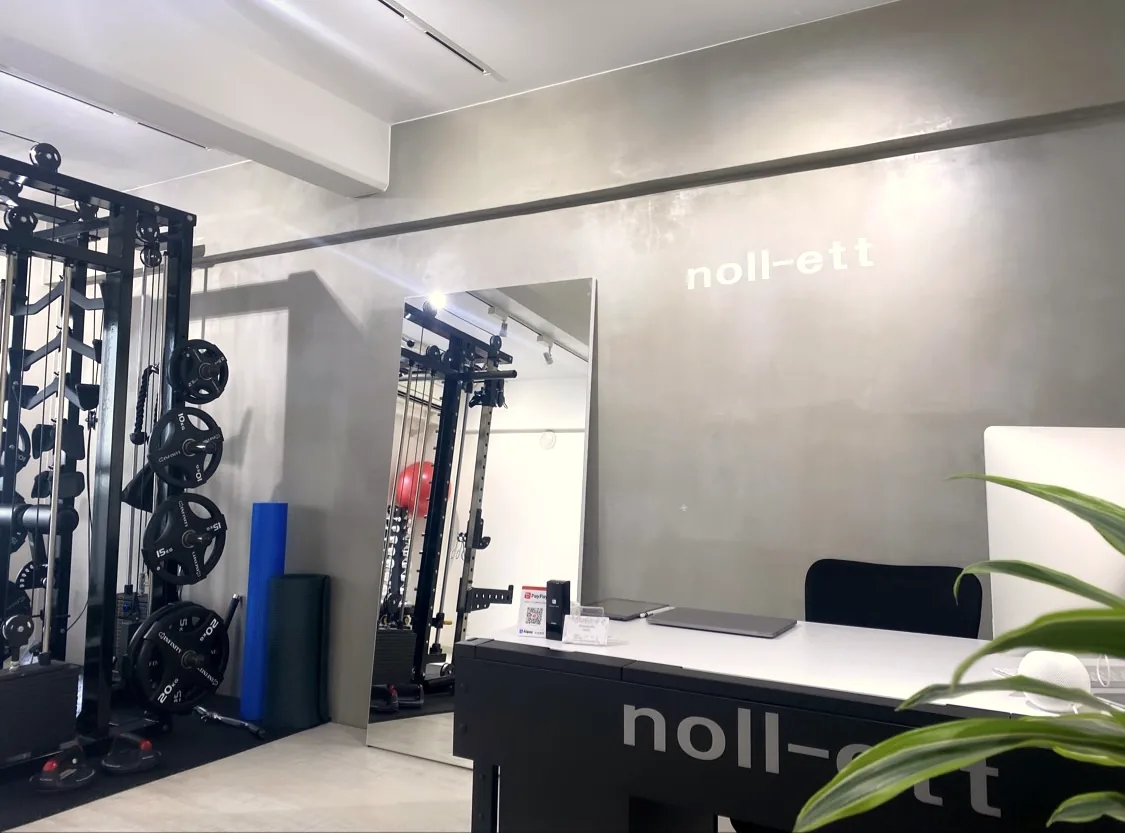 白・黒・グレーを基調にした完全個室。noll-ettのロゴが入った壁と受付カウンター、ケーブルマシンとプレート｜パーソナルジムnoll-ett渋谷店