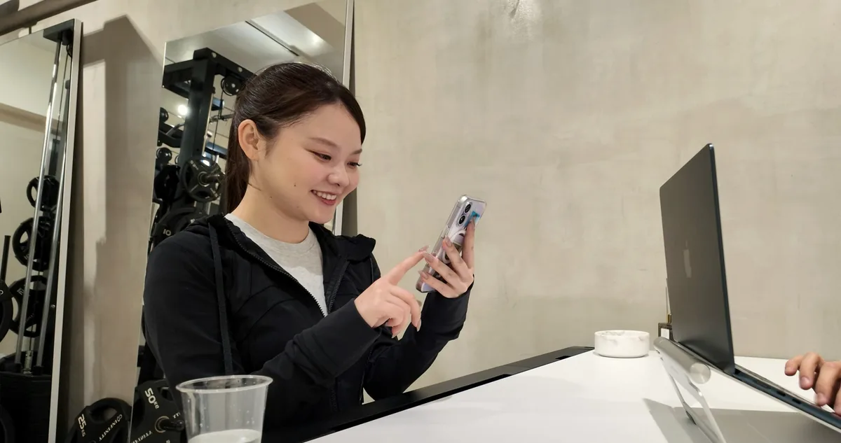 受付カウンターでスマートフォンを見ながら笑顔で話すお客様｜パーソナルジムnoll-ett渋谷店