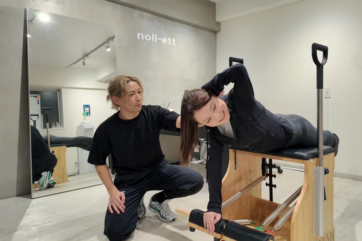 noll-ettのロゴがある完全個室で、ピラティスチェアを使って体側を伸ばすお客様とトレーナー｜パーソナルジムnoll-ett渋谷店